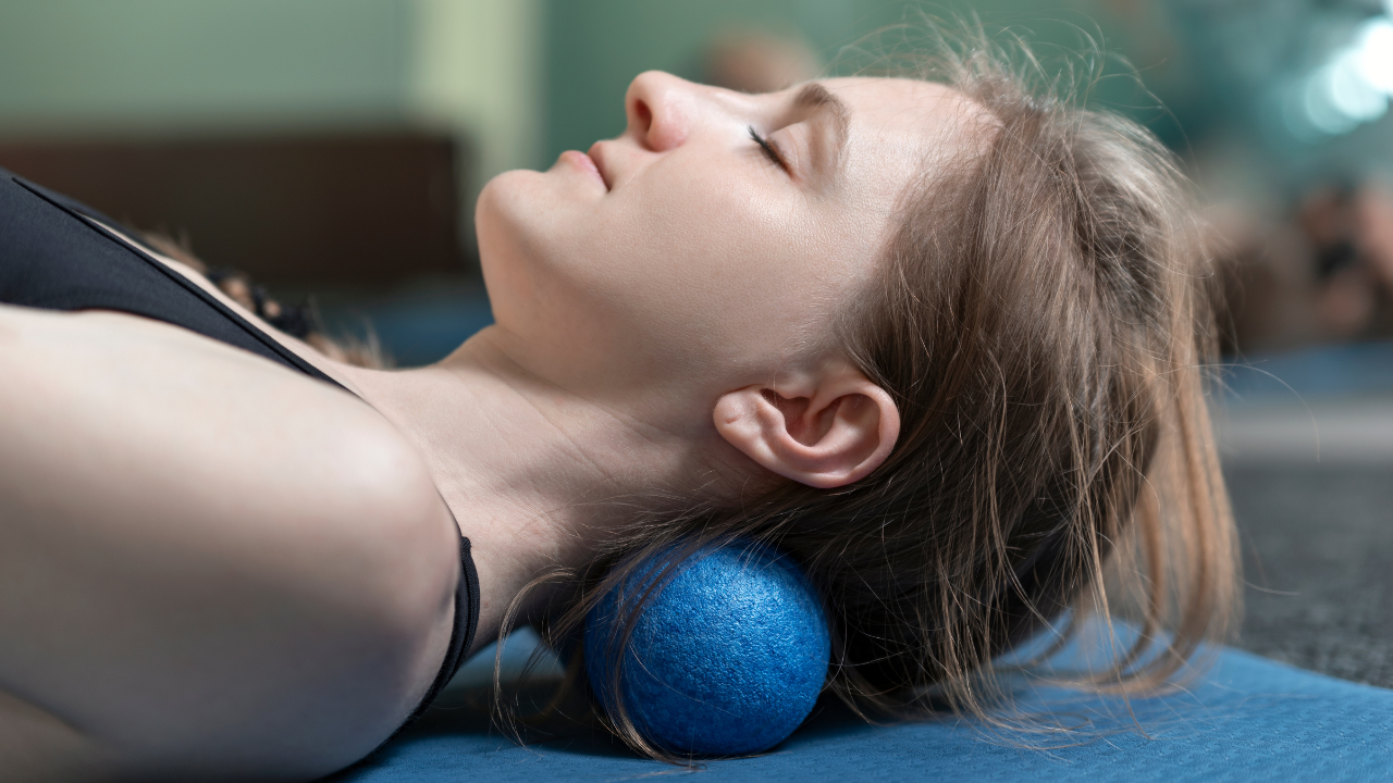 Frau in schwarzem Sporttop, die mit geschlossenen Augen auf dem Boden liegt und mit dem Nacken über einen Faszienball rollt